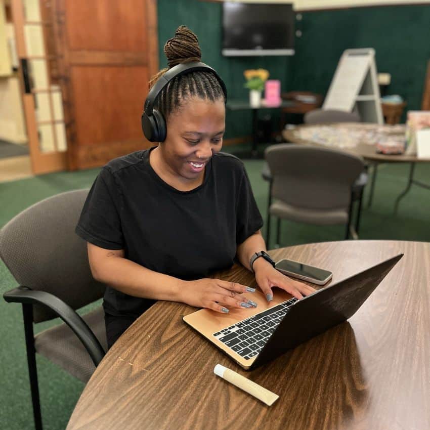 Woman writer sits cross-legged in a comfy chair with her headphones on writing